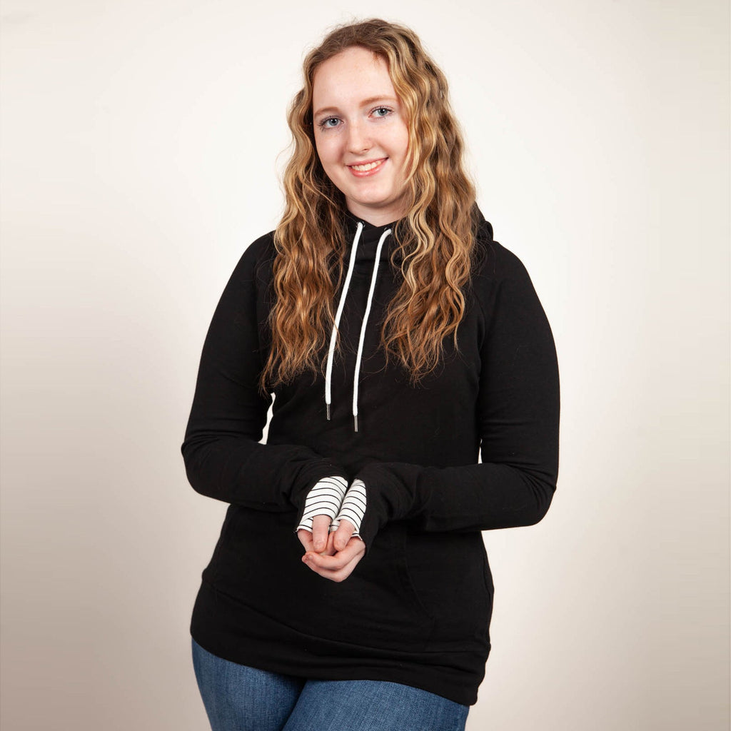 A woman wearing a black hoodie with thumbholes, watch cuff, a kangaroo pocket, and black and white pinstripe-lined hood. shown in size medium