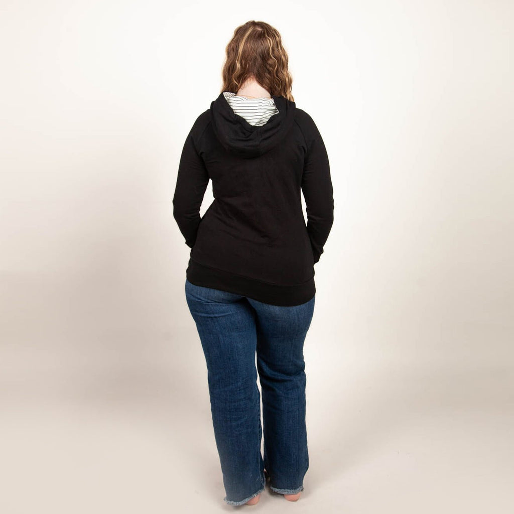 A woman wearing a black hoodie with thumbholes, watch cuff, a kangaroo pocket, and black and white pinstripe-lined hood. shown in size medium
