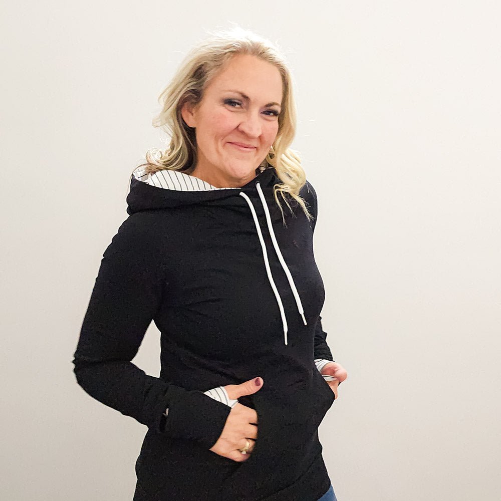 A woman wearing a black hoodie with thumbholes, watch cuff, a kangaroo pocket, and black and white pinstripe-lined hood. shown in size medium