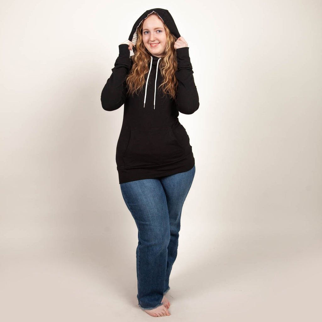 A woman wearing a black hoodie with thumbholes, watch cuff, a kangaroo pocket, and black and white pinstripe-lined hood. shown in size medium