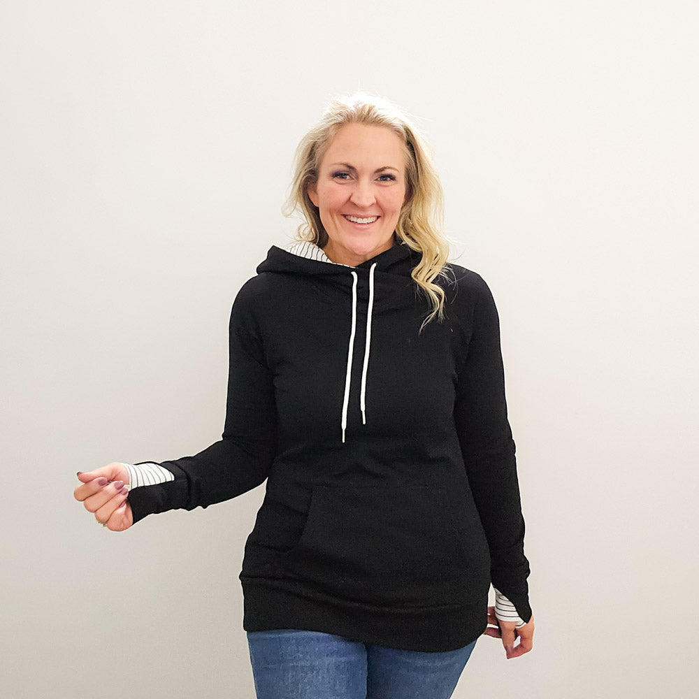 A woman wearing a black hoodie with thumbholes, watch cuff, a kangaroo pocket, and black and white pinstripe-lined hood. shown in size medium