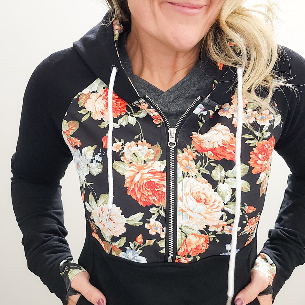 A woman wearing a black hoodie with a floral print on the front, featuring a half zip, floral print-lined hood and thumbholes, and kangaroo pocket. shown in size medium