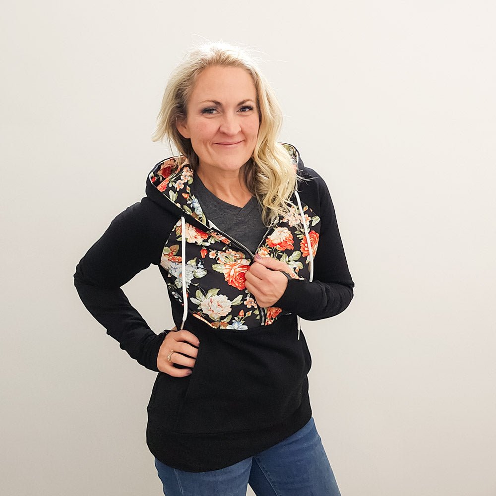 A woman wearing a black hoodie with a floral print on the front, featuring a half zip, floral print-lined hood and thumbholes, and kangaroo pocket. shown in size medium