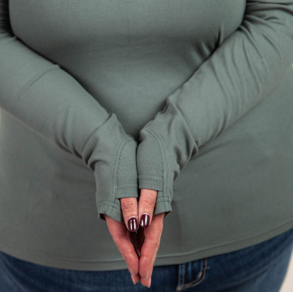A woman wearing a dark sage long sleeve, semi-fitted, henley top with thumbholes. 