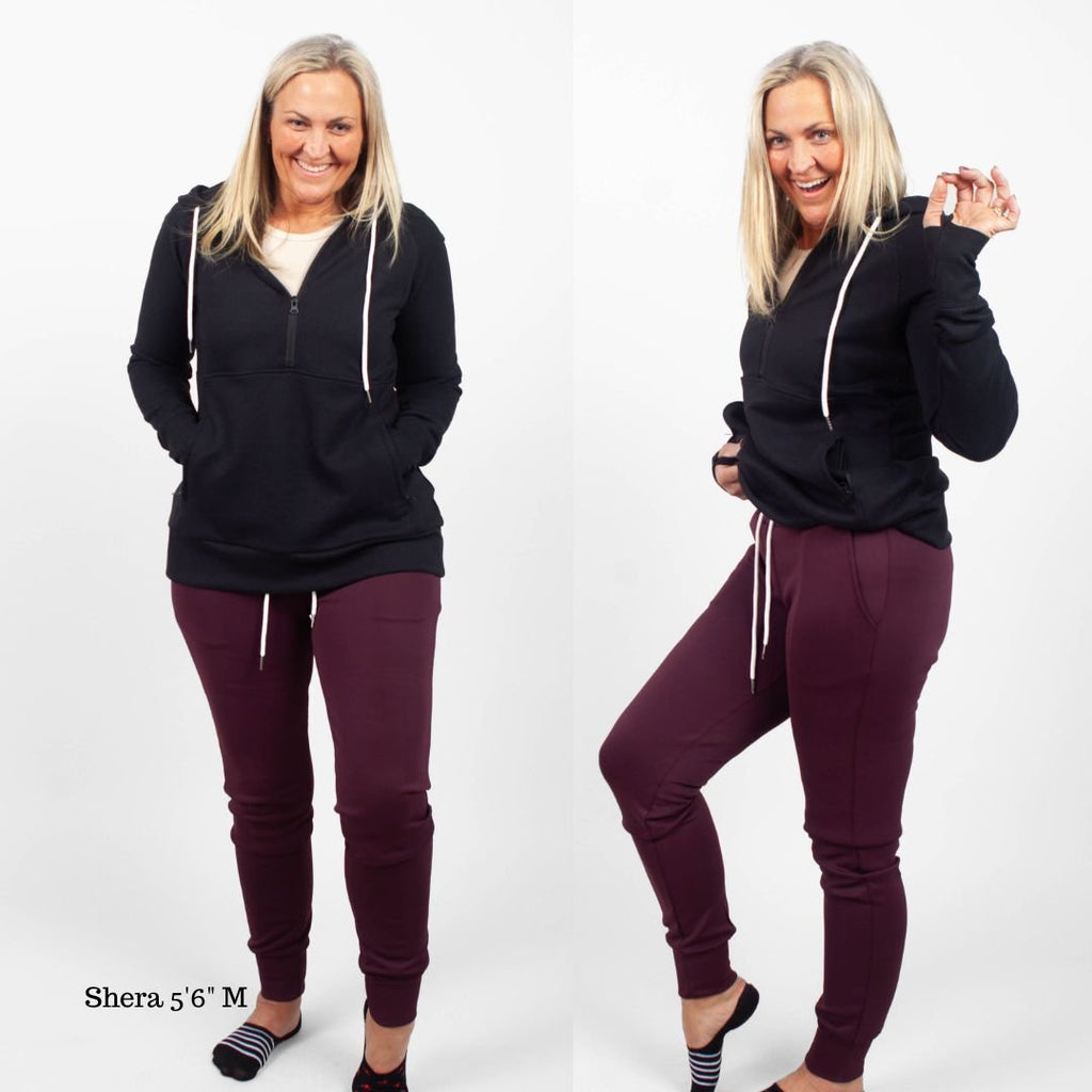 A woman standing, wearing  Eggplant Scuba Joggers 31" Inseam with a wide elastic waistband and tapered ankle cuffs, shown in size medium womens. paired with an Eclipse Black Scuba Half Zip hoodie featuring hood, kangaroo zipper pocket, and thumbholes shown in size medium. 