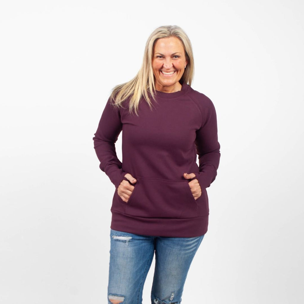 A woman wearing an eggplant purple crew neck sweatshirt with long sleeves, thumbholes, and kangaroo pocket. shown in size medium
