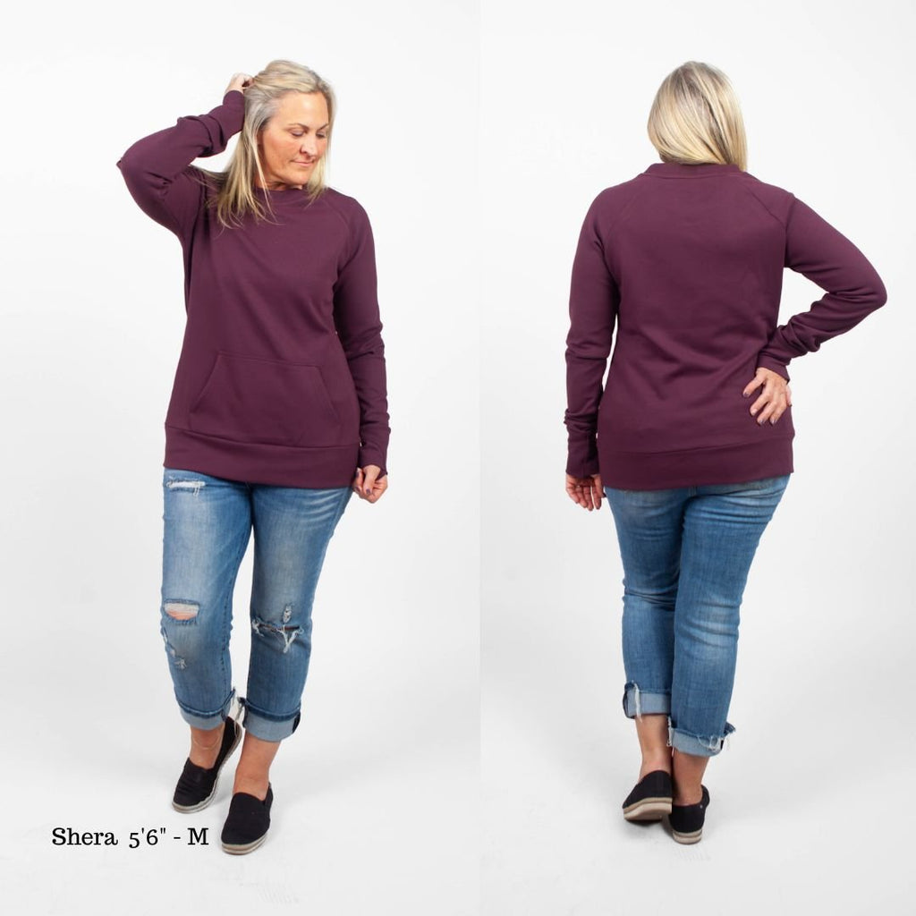 A woman wearing an eggplant purple crew neck sweatshirt with long sleeves, thumbholes, and kangaroo pocket. shown in size medium