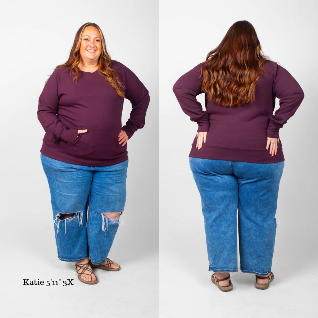 A woman wearing an eggplant purple crew neck sweatshirt with long sleeves, thumbholes, and kangaroo pocket. shown in size 3XL