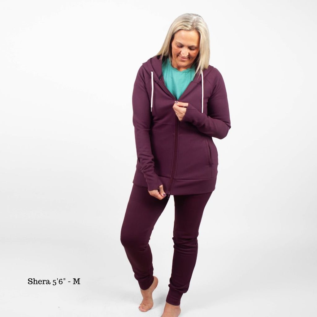 A woman standing, wearing  Eggplant Scuba Joggers 31" Inseam with a wide elastic waistband and tapered ankle cuffs, shown in size medium womens. paired with an Eggplant Scuba Full Zip  hoodie with a hood, featuring zipper pockets, and thumbholes. shown in size medium.