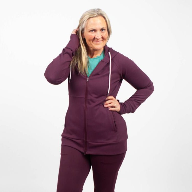 A woman wearing an eggplant purple full zip hoodie with a hood, featuring zipper pockets, and thumbholes. shown in size medium.
