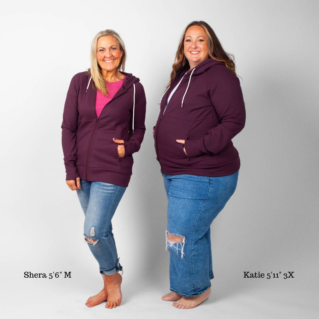 A woman wearing an eggplant purple full zip hoodie with a hood, featuring zipper pockets, and thumbholes. shown in size medium and 3XL