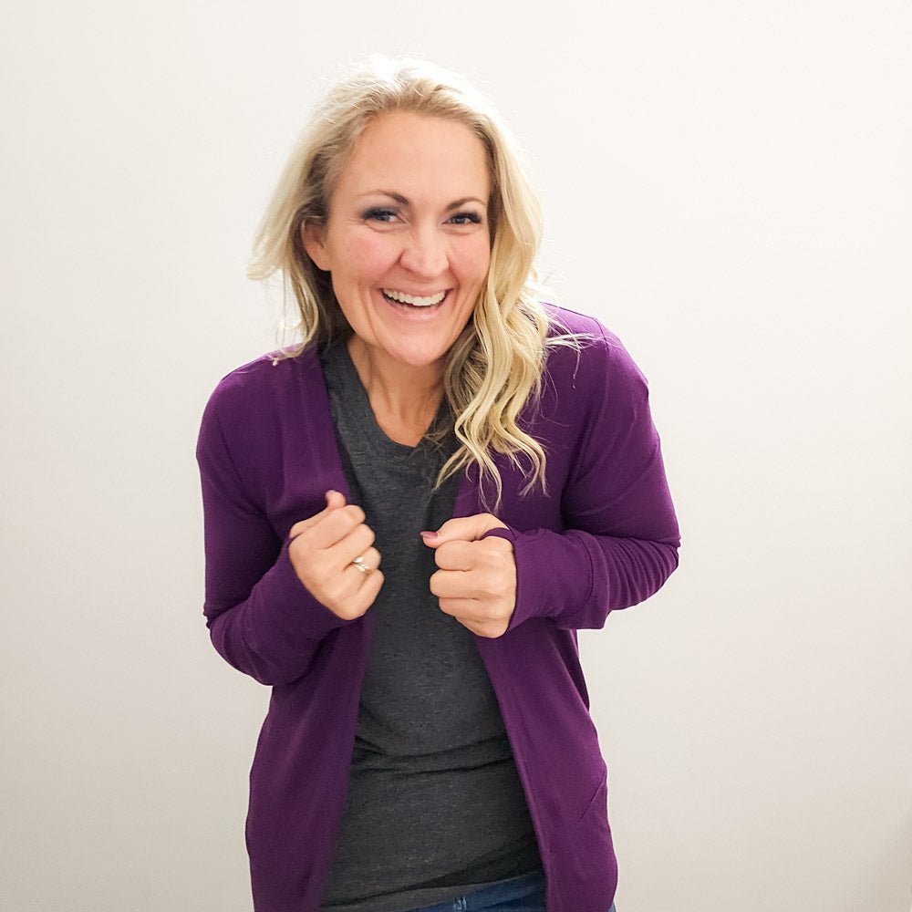 A woman wearing a purple open cardigan with long sleeves and thumbholes - shown in size medium. paired with a grey top and blue jeans.