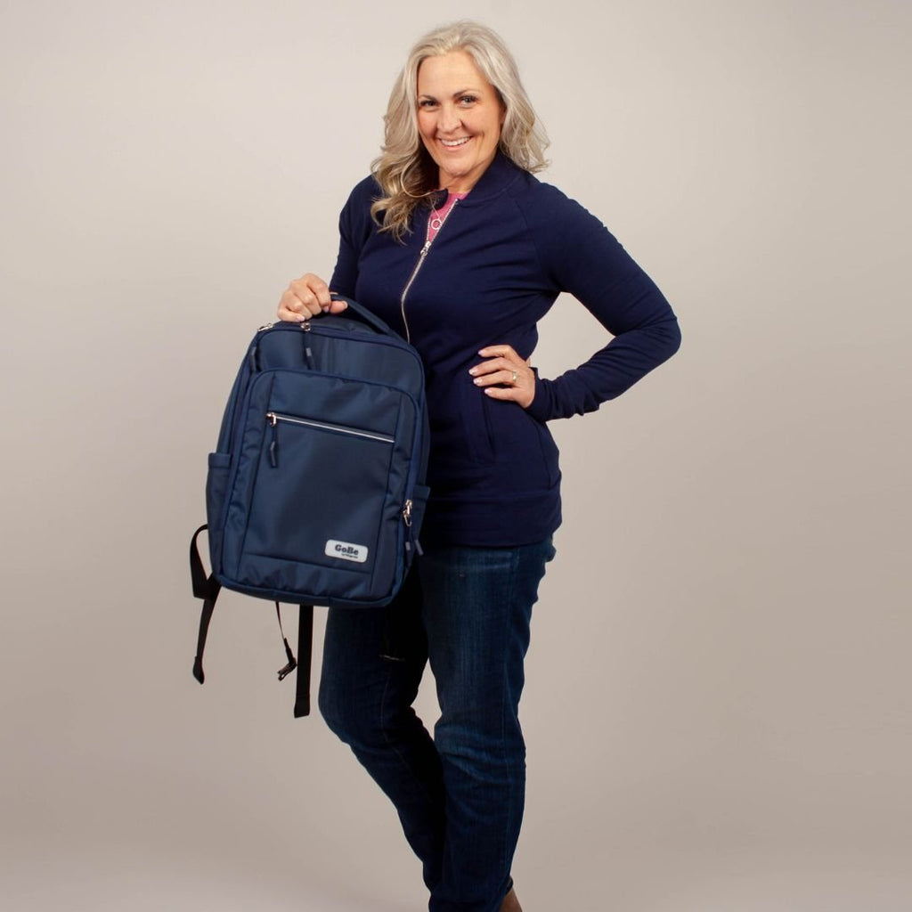 Go-Be by 7Degrees Backpack Navy  - stylish and functional travel bag for women. Lightweight and easy to carry with top handle. Woman with navy blue hoodie holding navy blue backpack by top handle up to waist.