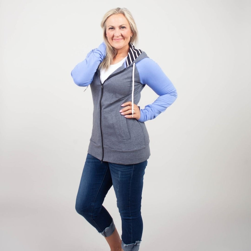 Woman wearing a gray and blue hoodie with side pockets, and a black and white striped pattern on the hood and thumbholes. shown in size medium