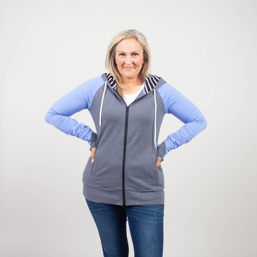 Woman wearing a gray and blue hoodie with side pockets, and a black and white striped pattern on the hood and thumbholes. shown in size medium