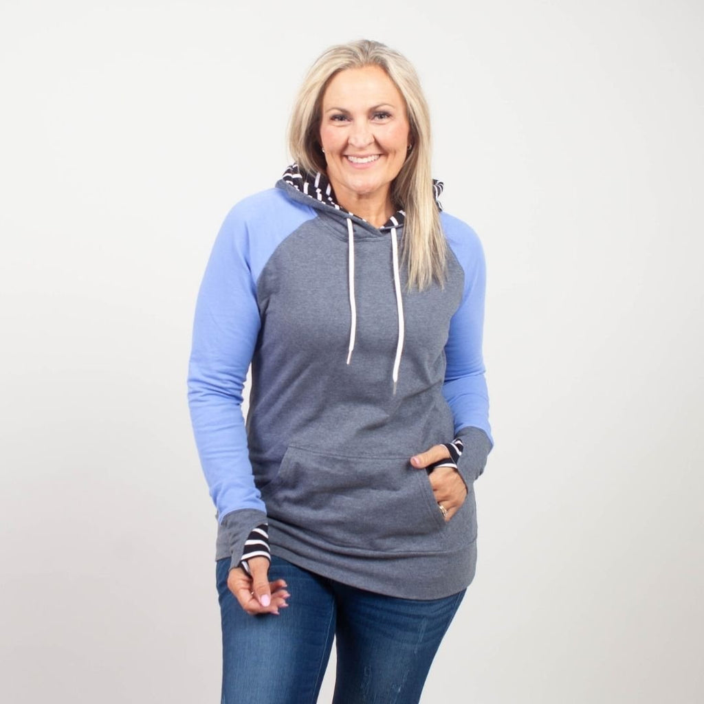 Woman wearing a gray and blue hoodie with kangaroo pocket, grey sleeve cuffs, and a black and white striped pattern on the hood and thumbholes. shown in size medium