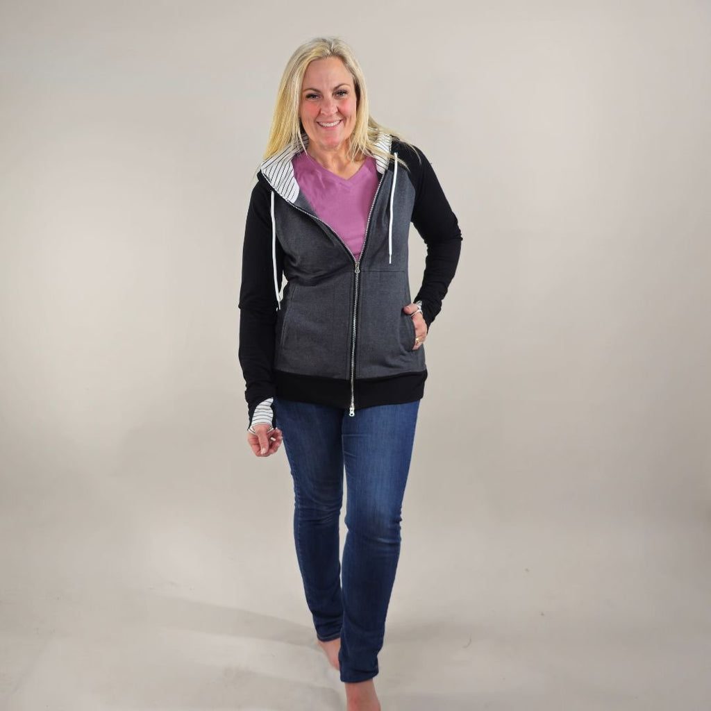 A woman wearing a dark gray hoodie with black sleeves and white pinstripes on the hood and thumb holes. shown in size medium