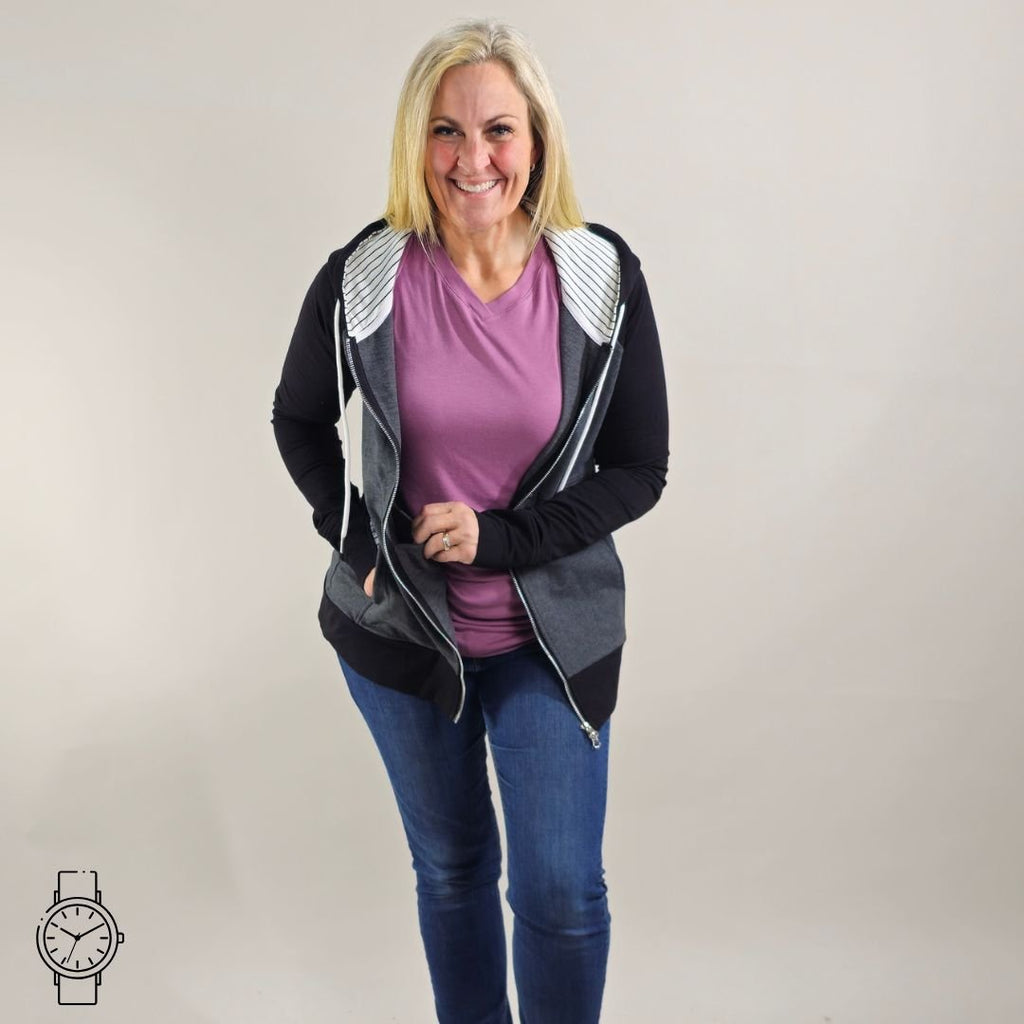 A woman wearing a dark gray hoodie with black sleeves and white pinstripes on the hood and thumb holes. shown in size medium