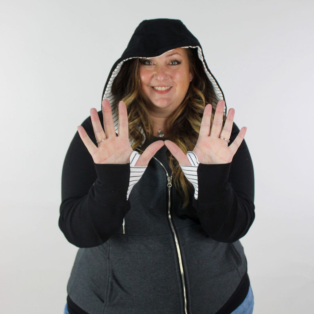 A woman wearing a dark gray hoodie with black sleeves and white pinstripes on the hood and thumb holes. shown in size 3XL