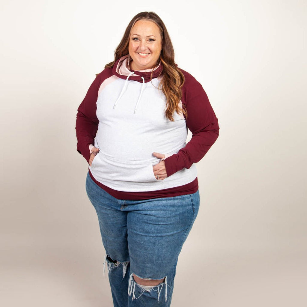 A woman wearing a two-tone hoodie with a burgundy color block design, featuring a hood, thumbholes, and a kangaroo pocket. shown in size 3XL