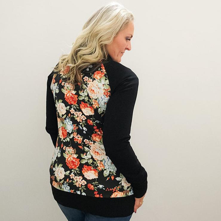 A woman wearing a floral print hoodless jacket with a stand collar, full front zipper, black sleeves, and floral  thumbholes. shown in size medium