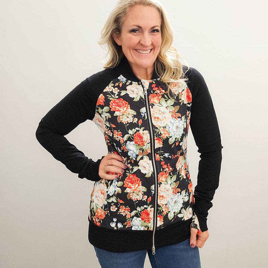 A woman wearing a floral print hoodless jacket with a stand collar, full front zipper, black sleeves, and floral  thumbholes. shown in size medium