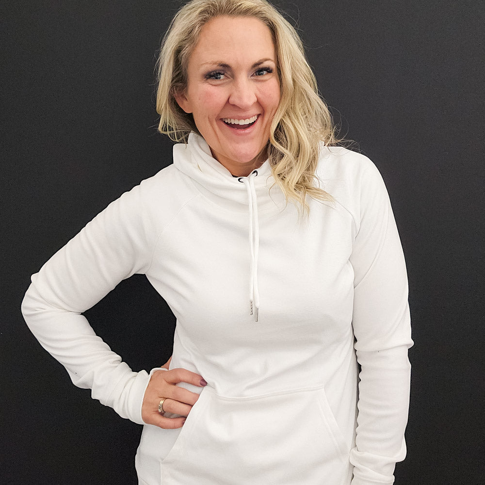 A woman wearing a white pullover hoodie with a hood, featuring a kangaroo front pocket and thumbholes on the sleeves. shown in size medium