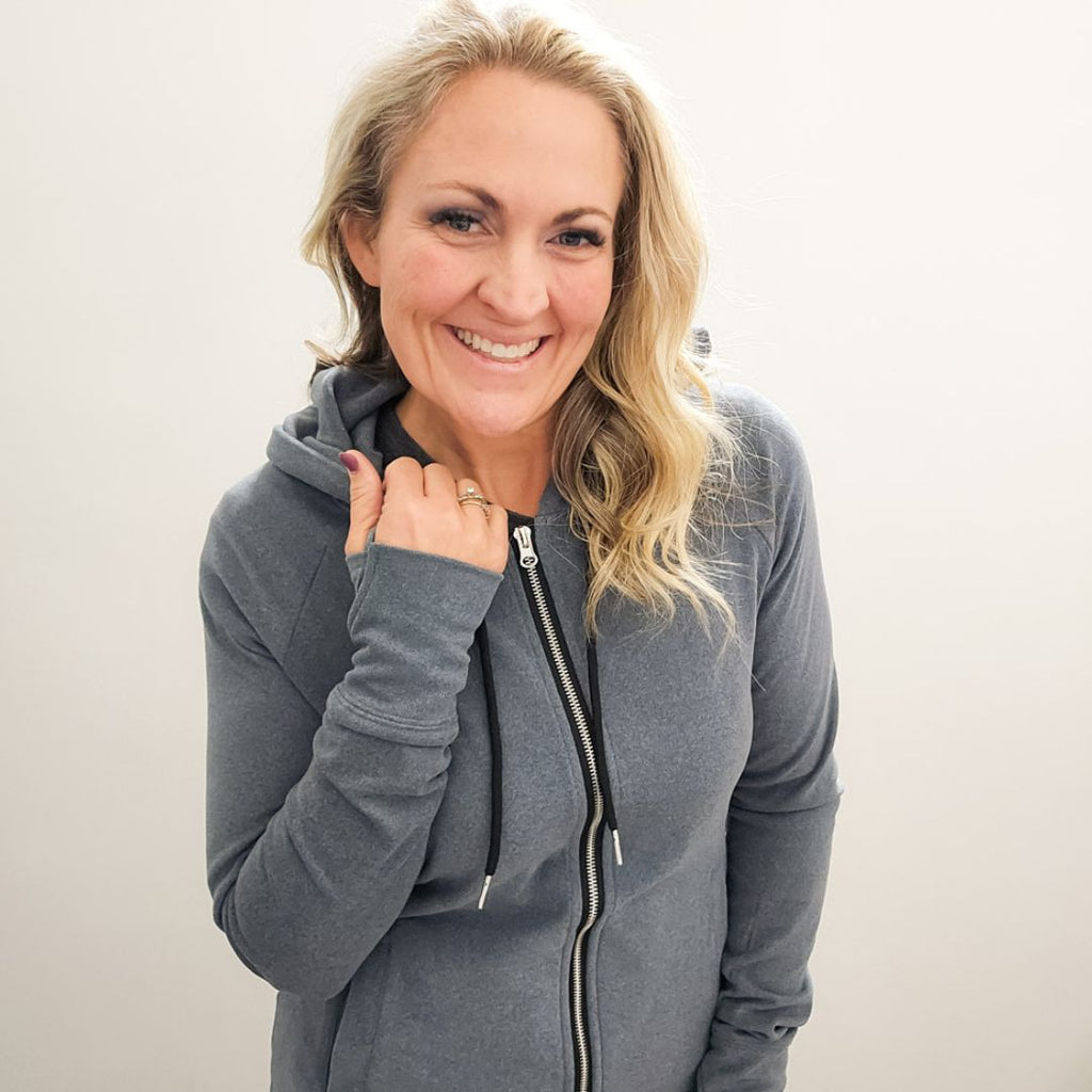 A woman wearing a grey zip-up hoodie with a hood, featuring a double zipper and thumbholes on the sleeves. shown in size medium