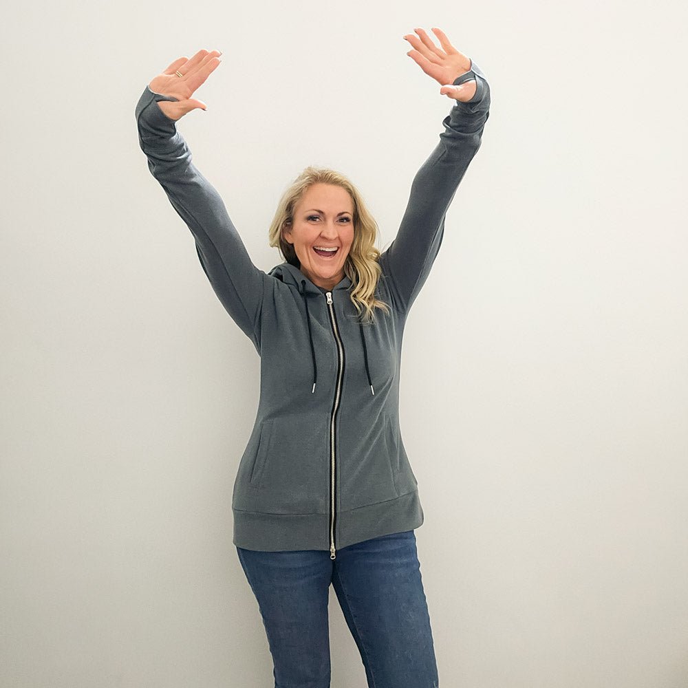 A woman wearing a grey zip-up hoodie with a hood, featuring a double zipper and thumbholes on the sleeves. shown in size medium