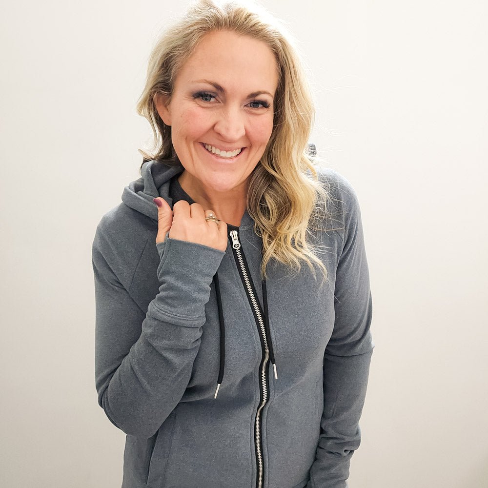 A woman wearing a grey zip-up hoodie with a hood, featuring a double zipper and thumbholes on the sleeves. shown in size medium