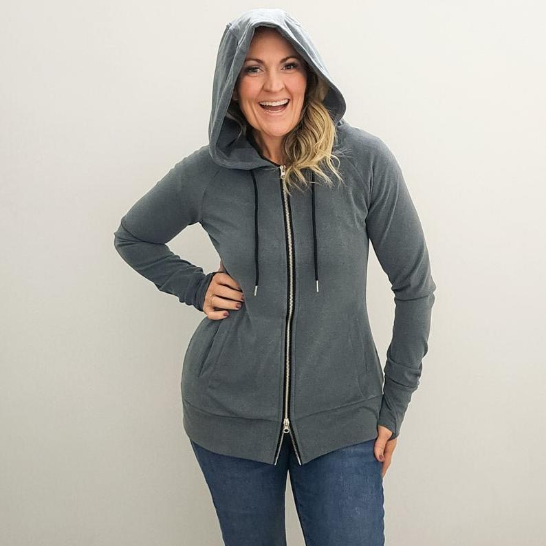 A woman wearing a grey zip-up hoodie with a hood, featuring a double zipper and thumbholes on the sleeves. shown in size medium