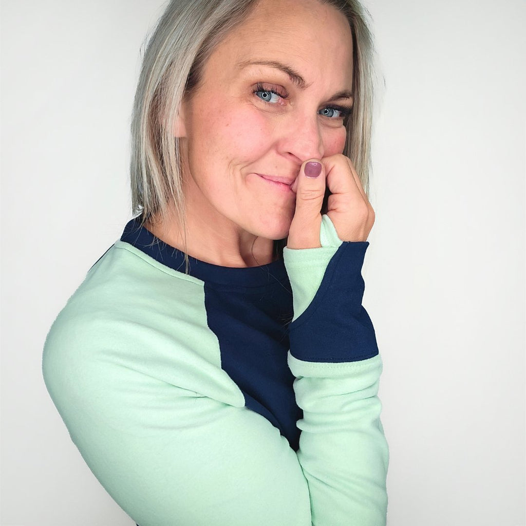 A woman wearing a long-sleeved crew neck pullover with a kangaroo pocket, in blue and green colors, with thumbholes. shown in size medium