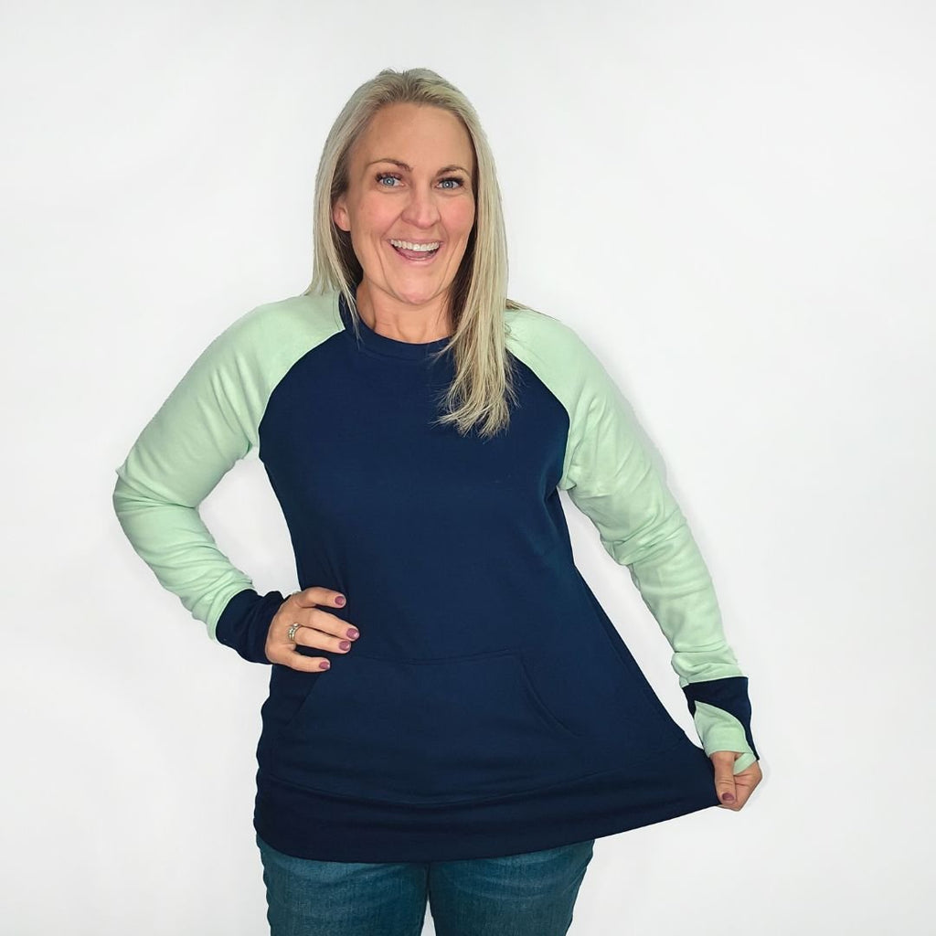 A woman wearing a long-sleeved crew neck pullover with a kangaroo pocket, in blue and green colors, with thumbholes. shown in size medium