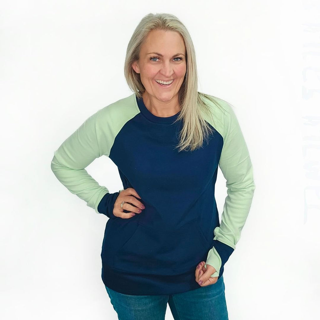 A woman wearing a long-sleeved crew neck pullover with a kangaroo pocket, in blue and green colors, with thumbholes. shown in size medium