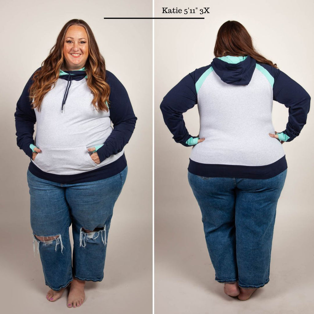 A woman wearing a color-blocked pullover with a hood, predominantly in navy and light heather gray with mint green accents. Features kangaroo pocket and thumbholes. Shown in size 3XL