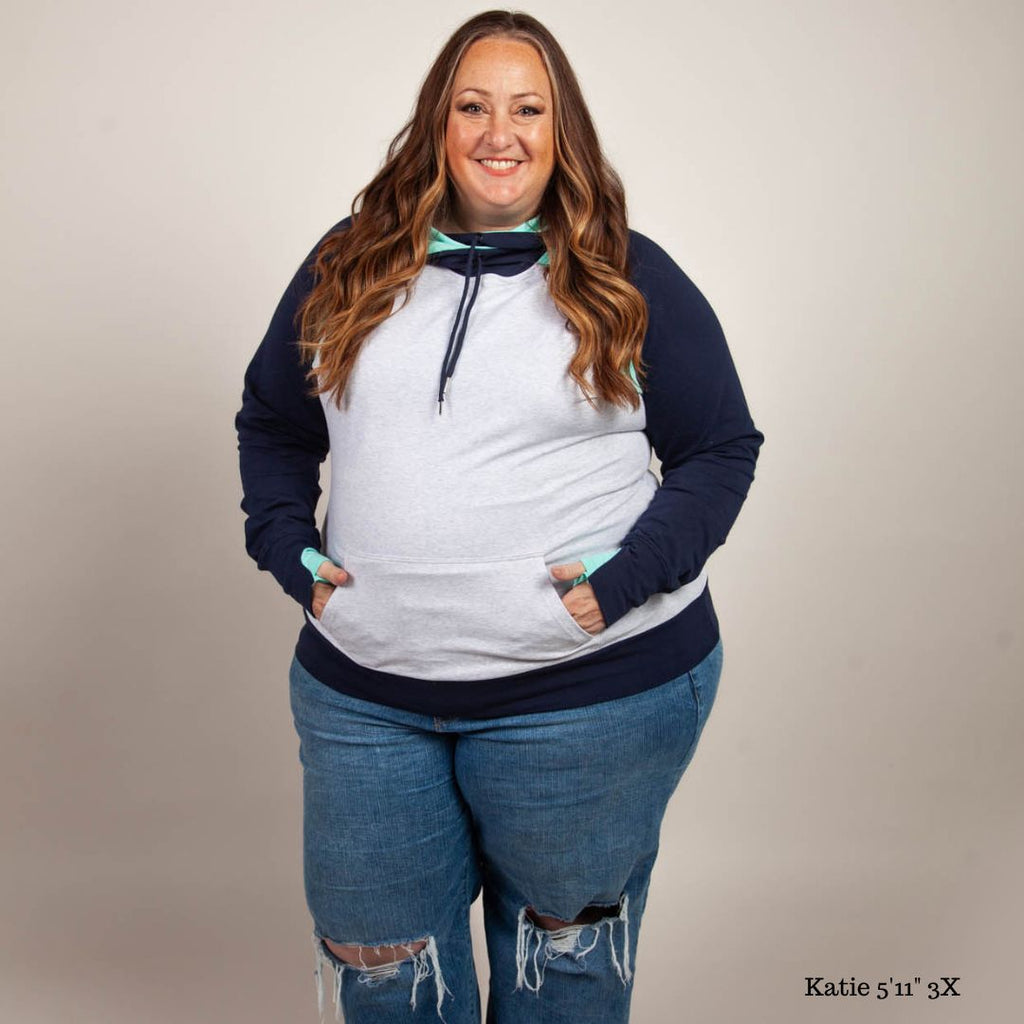 A woman wearing a color-blocked pullover with a hood, predominantly in navy and light heather gray with mint green accents. Features kangaroo pocket and thumbholes. Shown in size 3XL