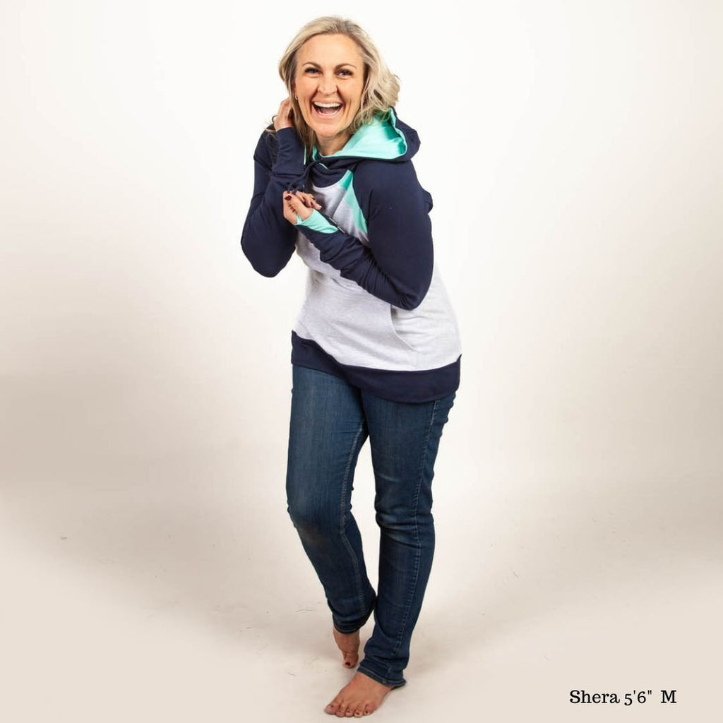 A woman wearing a color-blocked pullover with a hood, predominantly in navy and light heather gray with mint green accents. Features kangaroo pocket and thumbholes. Shown in size medium