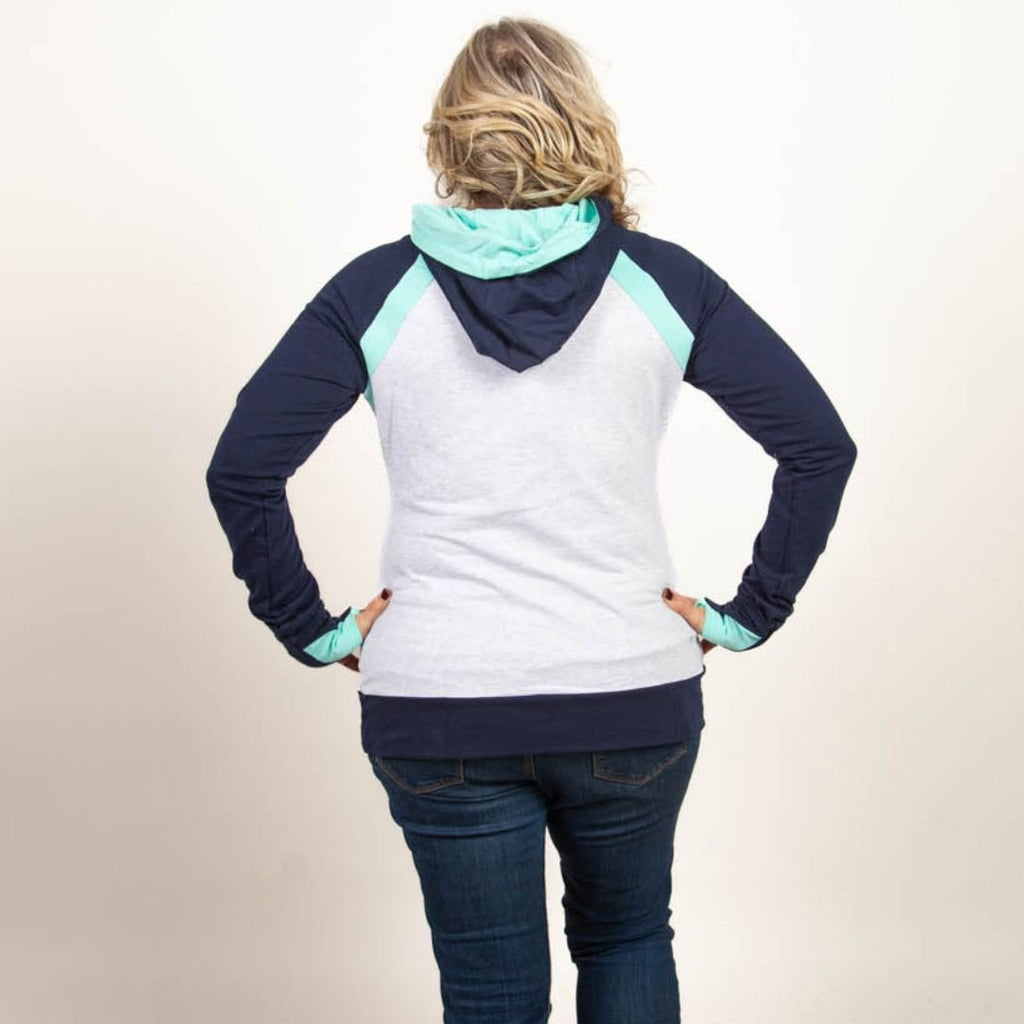 A woman wearing a color-blocked pullover with a hood, predominantly in navy and light heather gray with mint green accents. Features kangaroo pocket and thumbholes. Shown in size medium