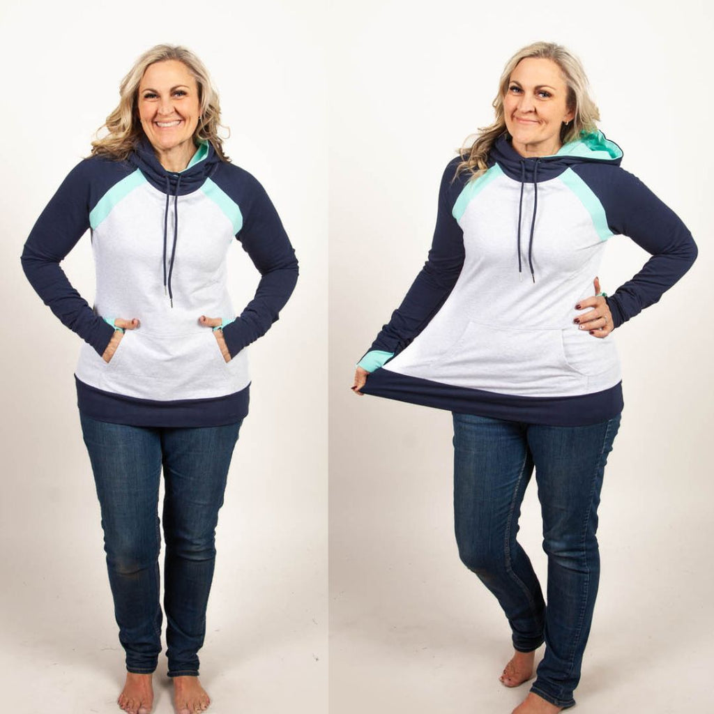 A woman wearing a color-blocked pullover with a hood, predominantly in navy and light heather gray with mint green accents. Features kangaroo pocket and thumbholes. Shown in size medium