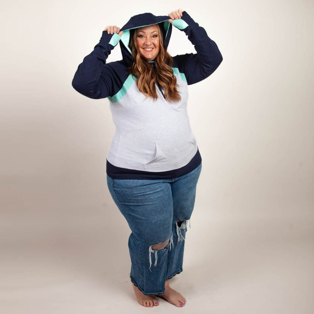 A woman wearing a color-blocked pullover with a hood, predominantly in navy and light heather gray with mint green accents. Features kangaroo pocket and thumbholes. Shown in size 3XL