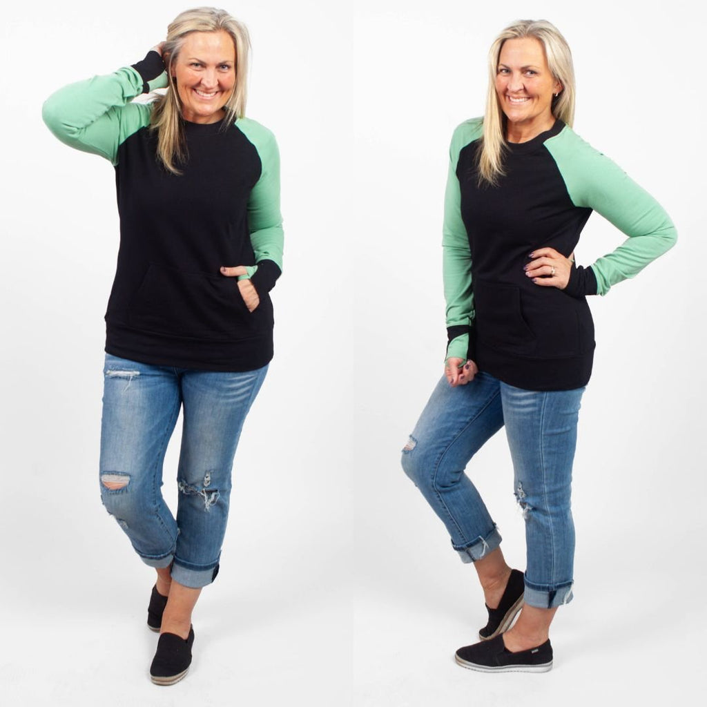 A woman wearing a long-sleeve crew neck pullover with green sleeves, a black base and kangaroo pocket. black sleeve cuffs and green accent on thumbhole. shown in size medium
