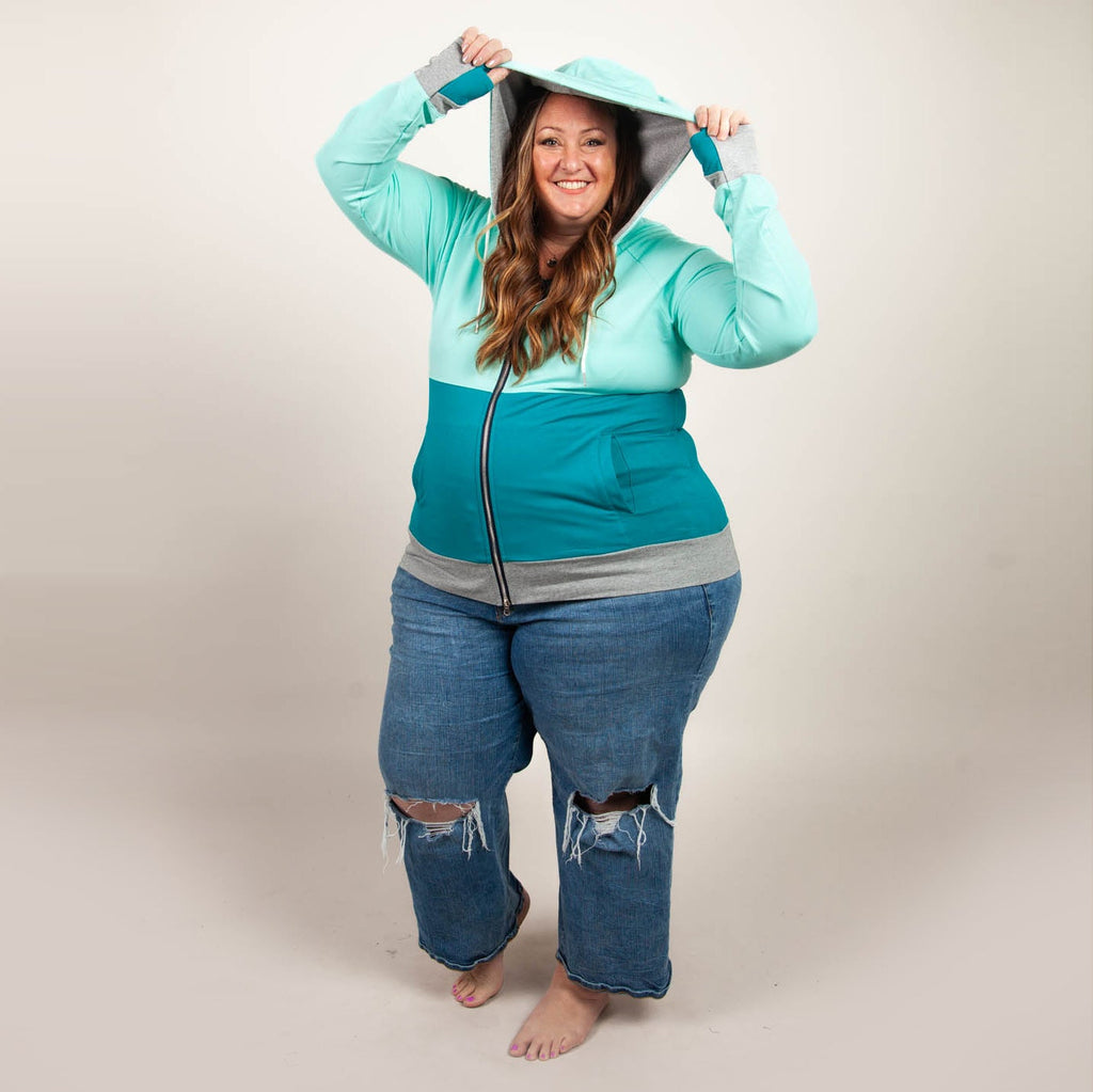 A woman wearing a color-block hoodie with mint green at the top, dark turquoise at the bottom, and heather gray accents. The hoodie has a full double zip, hood, thumbholes, and side pockets. shown in size 3XL