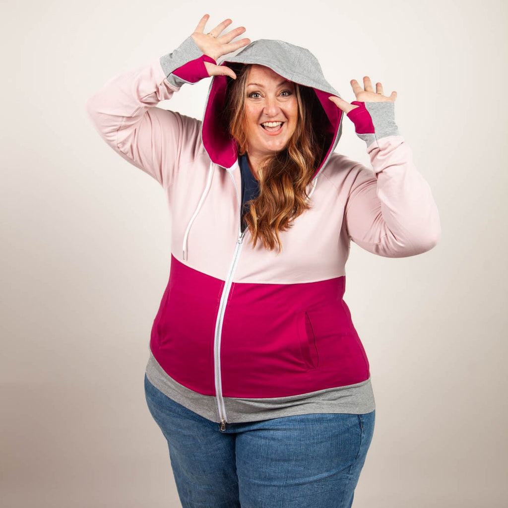 A woman wearing a color-blocked hoodie with light pink on the top, raspberry pink on the bottom, and heather gray accents. Features include side pockets, thumbholes, and raspberry pink lined hood. shown in size 3XL