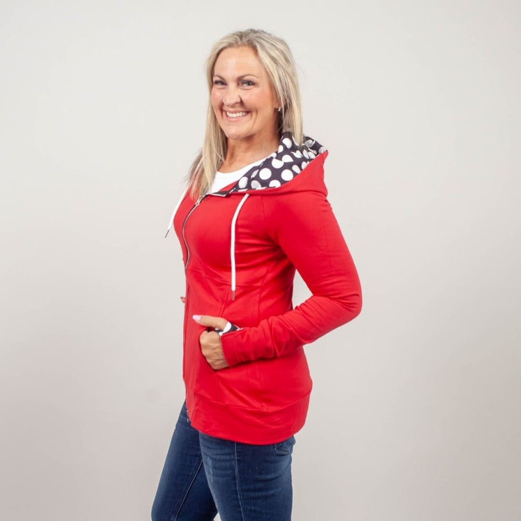 Woman wearing a red zip-up hoodie with a black and white dot lined hood, double zipper, and thumbholes. Shown in size medium