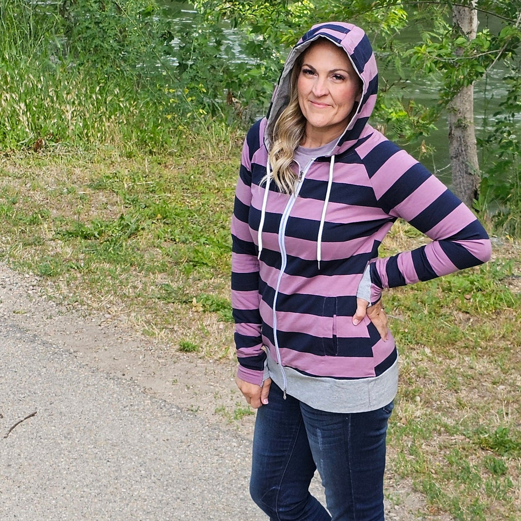 A woman wearing a striped hoodie with purple and navy colors, featuring a full zipper, hood, and thumbholes. shown in size medium