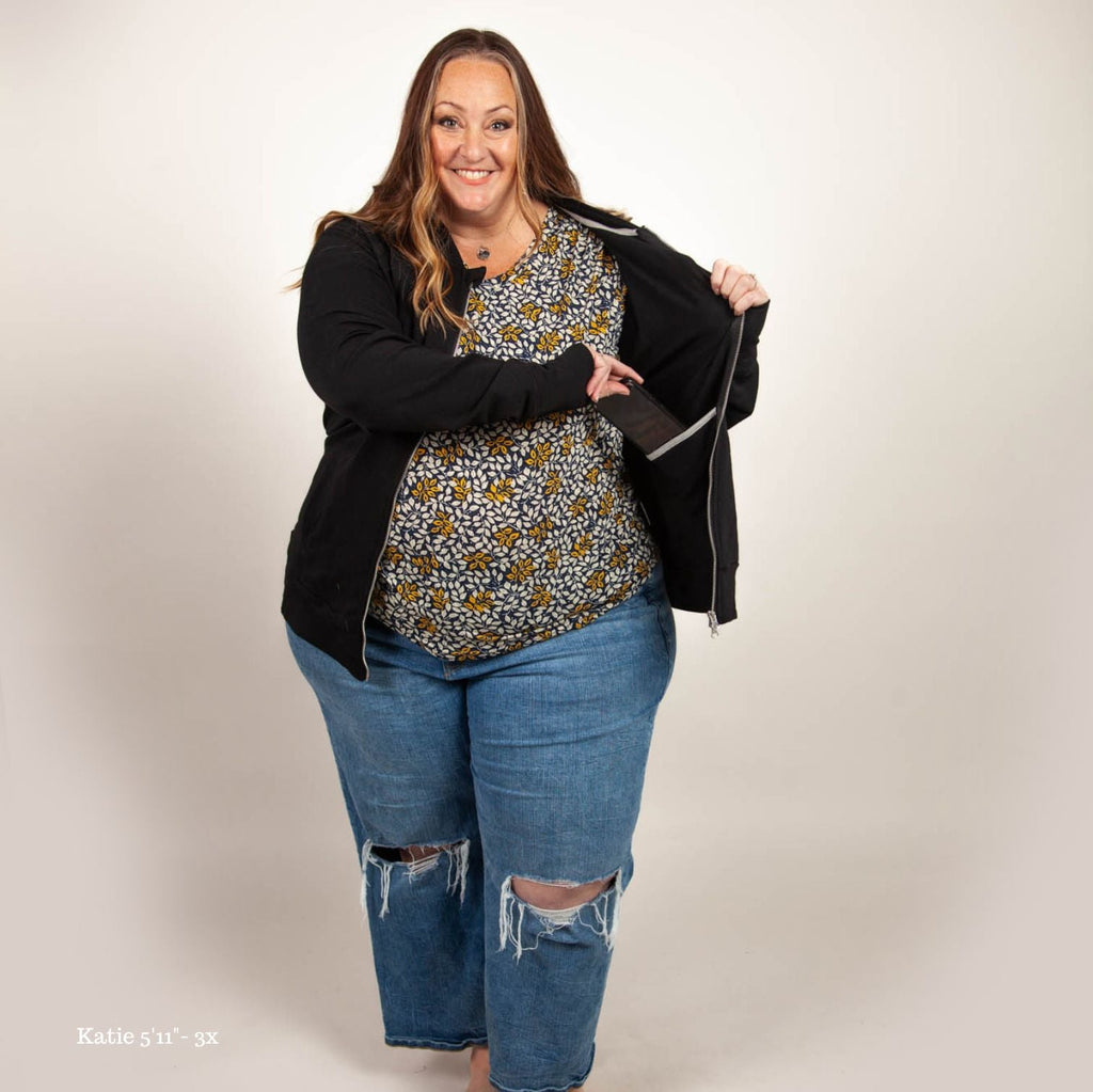A woman wearing a v-neck shirt with a floral pattern in yellow and white on a dark blue background. paired with black no-hood zip up jacket with a stand collar, full front zipper, and thumbholes.  shown in 3XL