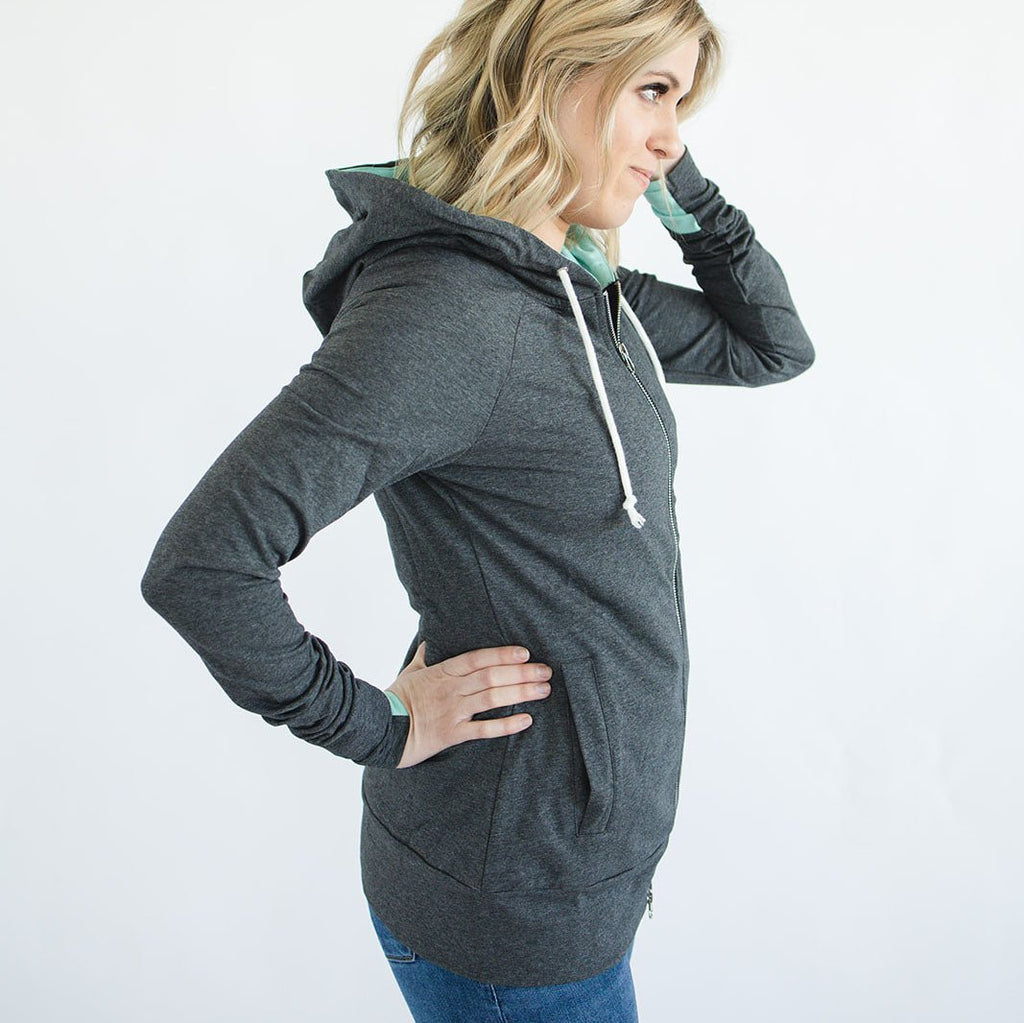 Woman wearing a charcoal grey full-zip hoodie with mint green accents. Features a deep lined hood, accent thumbholes without cuffs, longer body length, extra-long sleeves, large side welt pockets, and a two-way zipper. Made from a medium-weight cotton-spandex blend terry knit for a flattering hourglass fit Shown in Small Shop7degrees